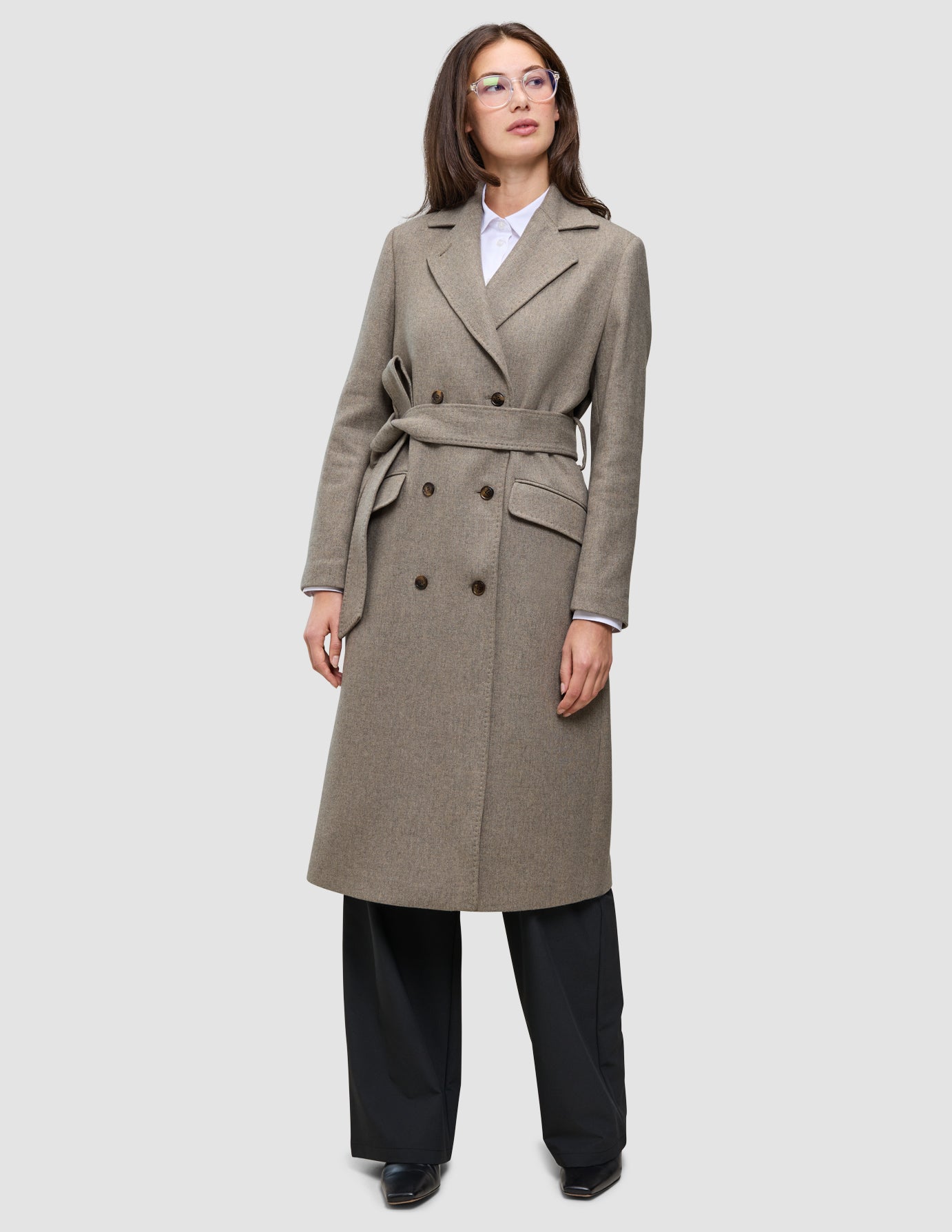 Eine Person, die einen Women Wool Coat Greige trägt – einen zweireihigen, knielangen, beigen Wollmantel mit einem passenden Bindegürtel –, dazu ein weißes Hemd und eine schwarze Hose, die das klare skandinavische Design unterstreichen.